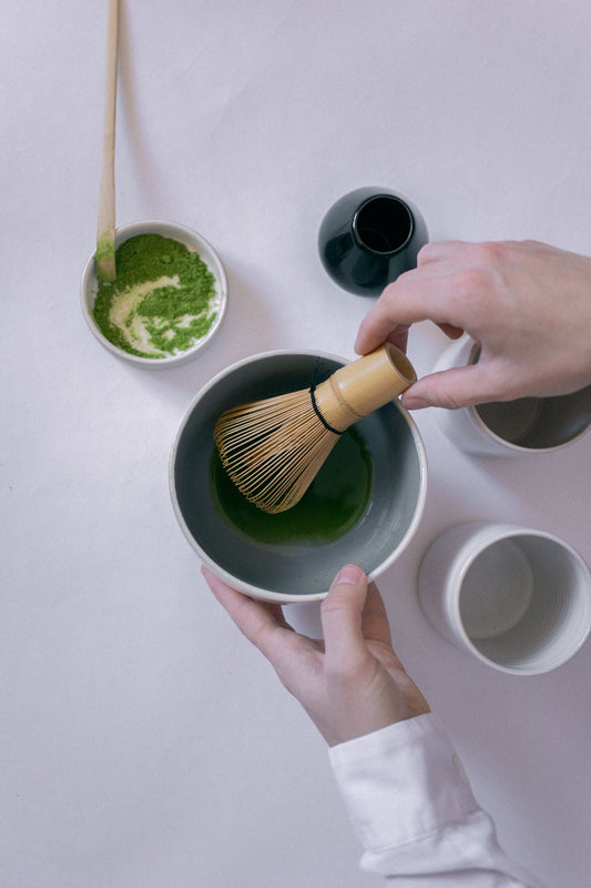 Hands preparing matcha with a bamboo whisk, ceramic bowl, and tea tools arranged on a light table