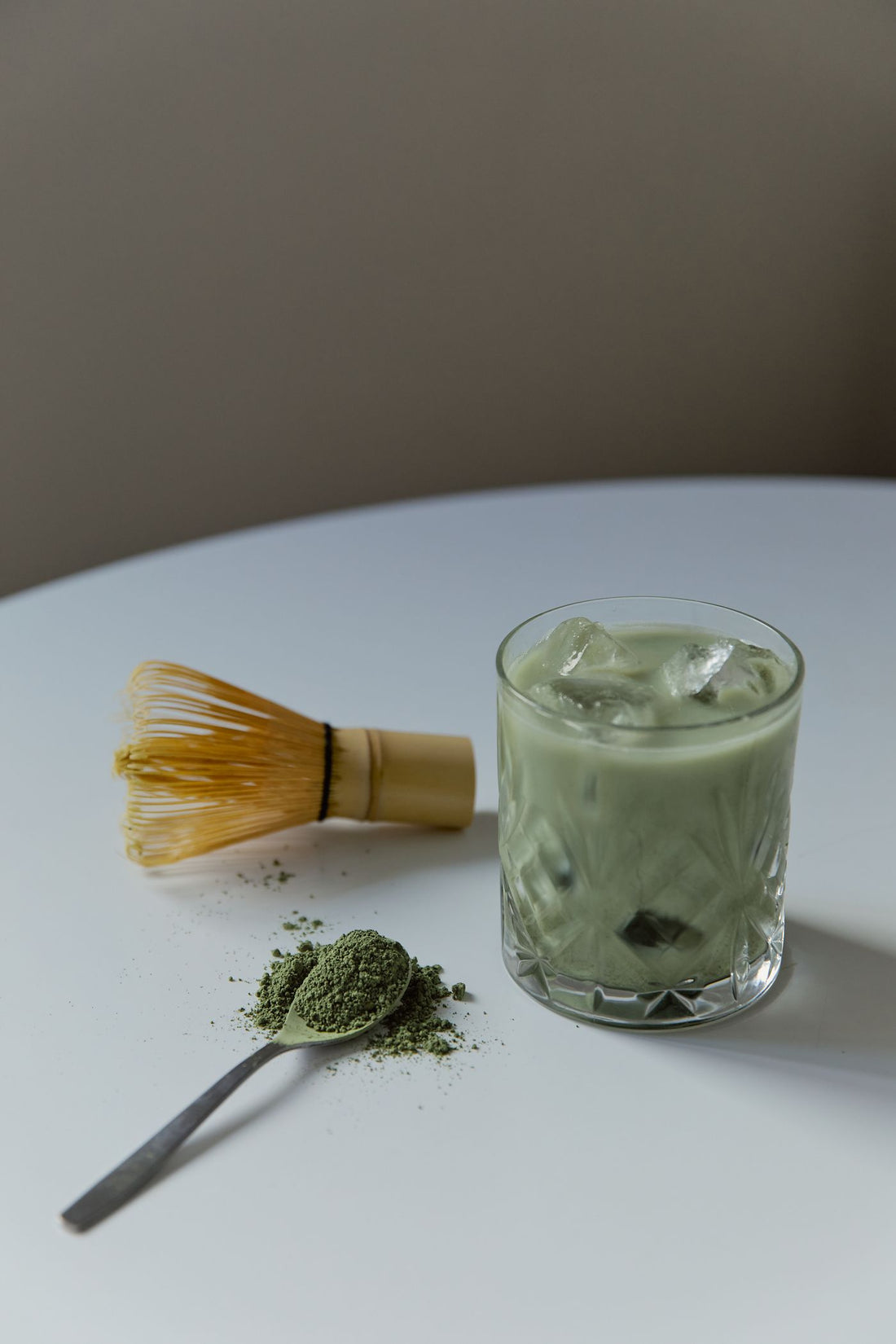 Iced matcha latte beside a bamboo whisk and powder on a pale table