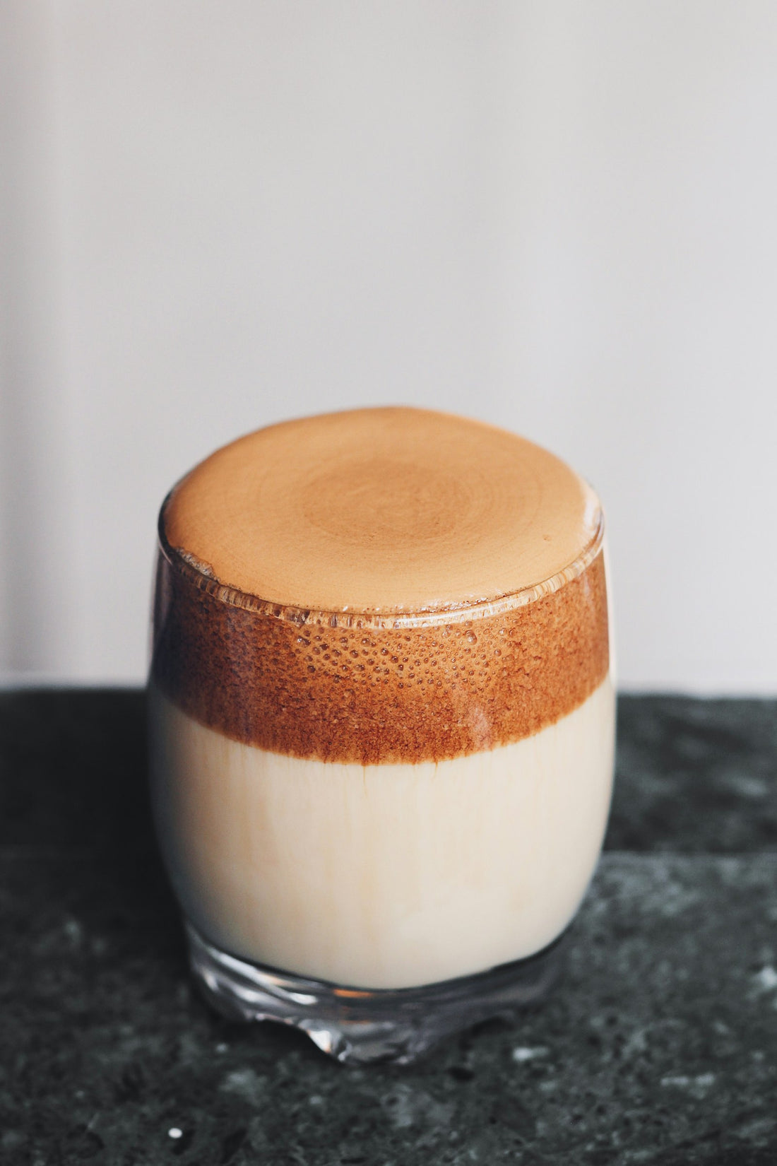 Layered tan latte with thick brown foam in a clear rounded glass